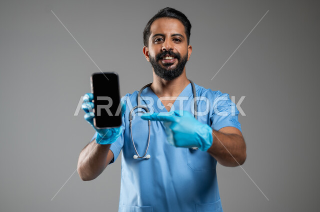 العمل في القطاع الصحي, دمج التكنولوجيا و التقنية في الحياة العملية, بورتريه مقرب لطبيب عربي سعودي خليجي يرتدي يونيفورم العمل يشير باصبع السبابة الى شاشة سوداء فارغة عبر الهاتف المحمول، النظر الى الكاميرا بتعابير السرور, خلفية رمادية، محتوى حساس، استخدام حساس