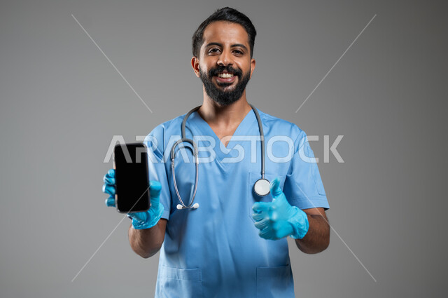 رفع اصبع الابهام للأعلى بتعابير علامة الجودة, دمج التكنولوجيا و التقنية في الحياة العملية, بورتريه مقرب لطبيب عربي سعودي خليجي يرتدي يونيفورم العمل يقوم بعرض شاشة فارغة سوداء عبر الهاتف المحمول، الشعور بالرضا و القبول, خلفية رمادية، محتوى حساس، استخدام حساس