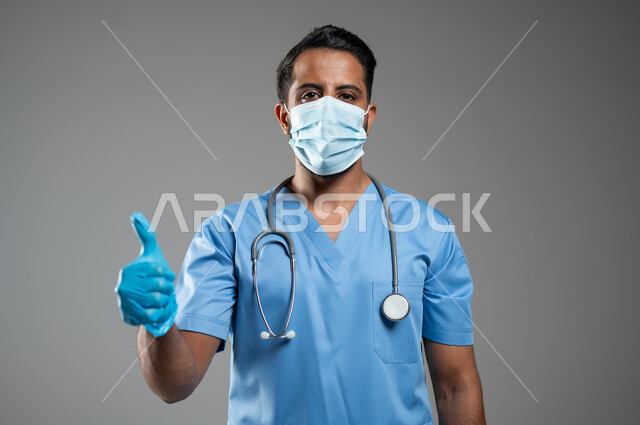 ارتداء الكمامه لتجنب انتشار فايروس كورونا, الرضا بجوده الخدمات الطبية، الالتزام بالاحترازات الوقائيه ضد كوفيد 19, بورتريه مقرب لممرض عربي سعودي خليجي يرتدي يونيفورم العمل يشير بإصبع الإبهام نحو الاعلى، خلفية رمادية، محتوى حساس، استخدام حساس