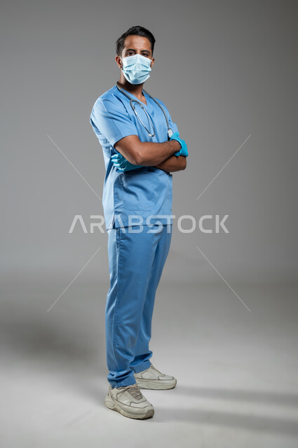 الالتزام بالاحترازات الوقائيه، تجنب انتشار كوفيد 19, بورتريه لممرض عربي خليجي سعودي يرتدي يونيفورم العمل يقف بتعابير ايدي متقاطعة وينظر الى الكاميرا ,ارتداء الكمامه للوقاية من فايروس كورونا، الجسم بالطول الكامل، خلفية رمادية, محتوى حساس, استخدام حساس