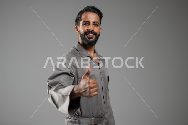 الشعور بالرضا و القبول والموافقة، النظر إلى الكاميرا بتعابير السرور، بورتريه مقرب لعامل صيانة عربي سعودي خليجي يرتدي يونيفورم العمل يرفع أصبع الابهام نحو الأعلى بإيماءات علامة الجودة، المهارة و اتقان العمل، خلفية رمادية، محتوى حساس، استخدام حساس