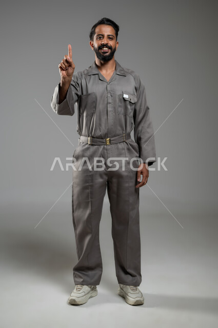 اشارة تمثل الرقم واحد، بورتريه لعامل صيانة عربي سعودي خليجي مبتسم يرتدي يونيفورم العمل يشير بأصبع السبابة نحو الأعلى، النظر الى الكاميرا بإيماءات السرور، المهارة و اتقان العمل،  الجسم بالطول الكامل، خلفية رمادية, محتوى حساس, استخدام حساس