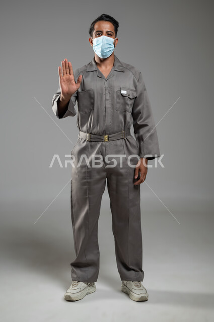 الالتزام بالاحترازات الوقائيه ضد كوفيد 19، رفع اليد للحذر من الاقتراب و طلب التوقف والابتعاد، التقيد بلبس الكمامه، بورتريه لعامل صيانة عربي سعودي خليجي يرفع يده ليتجنب المصافحة واقتراب الاخرين منه، الجسم بالطول الكامل، خلفية رمادية، استخدام حساس، محتوى حساس