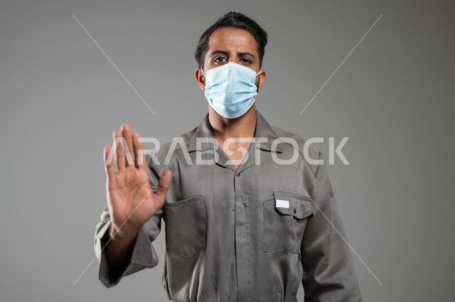 رفع اليد للحذر من الاقتراب و طلب التوقف والابتعاد، الالتزام بالاحترازات الوقائيه ضد كوفيد 19، التقيد بلبس الكمامه، بورتريه مقرب لعامل صيانة عربي سعودي خليجي ينظر إلى الكاميرا يرفع يده ليتجنب المصافحة واقتراب الاخرين منه، خلفية رمادية، استخدام حساس، محتوى حساس