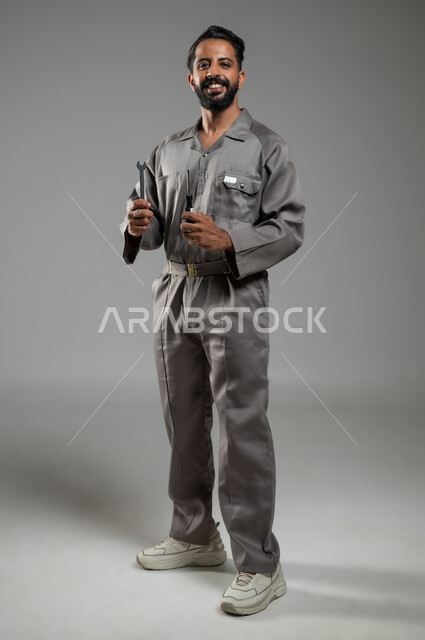 Using maintenance tools and equipment, a smiling repair technician looks at the camera with gestures of pleasure, skill and mastery of work, portrait of a Saudi Arabian Gulf maintenance worker wearing a work uniform holding a wrench and a screwdriver, full-length body, gray background, sensitive content, sensitive use