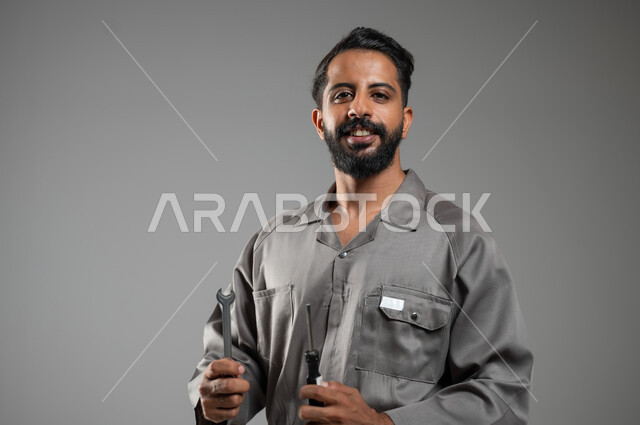 فني تصليح مبتسم ينظر إلى الكاميرا بإيماءات السرور، المهارة و اتقان العمل، استخدام أدوات ومعدات الصيانة، بورتريه مقرب لعامل صيانة عربي سعودي خليجي يرتدي يونيفورم العمل يحمل مفتاح الربط و مفك البراغي، خلفية رمادية, محتوى حساس, استخدام حساس