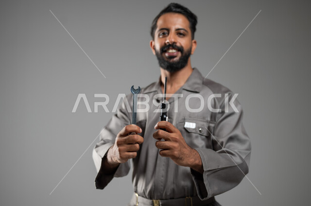 فني تصليح مبتسم ينظر إلى الكاميرا بإيماءات السرور، المهارة و اتقان العمل، استخدام أدوات ومعدات الصيانة، بورتريه مقرب لعامل صيانة عربي سعودي خليجي يرتدي يونيفورم العمل يحمل مفتاح الربط ومفك البراغي، خلفية رمادية, محتوى حساس, استخدام حساس