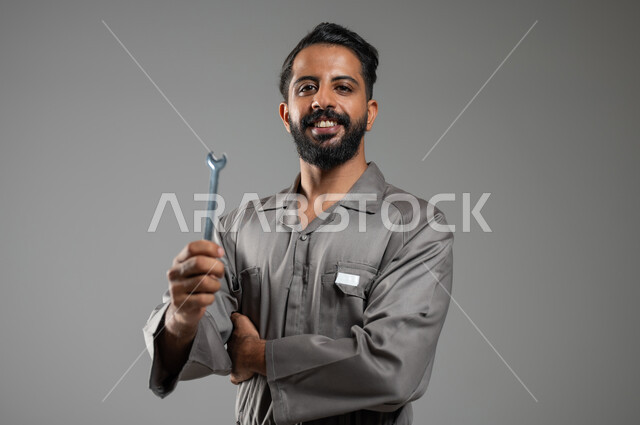 المهارة و اتقان العمل، بوتريه مقرب لعامل صيانة يرتدي يونيفورم العمل يحمل بيده مفتاح الربط، فني تصليح مبتسم ينظر إلى الكاميرا بإيماءات السرور، إستخدام أدوات ومعدات الصيانة، خلفية رمادية, محتوى حساس, استخدام حساس