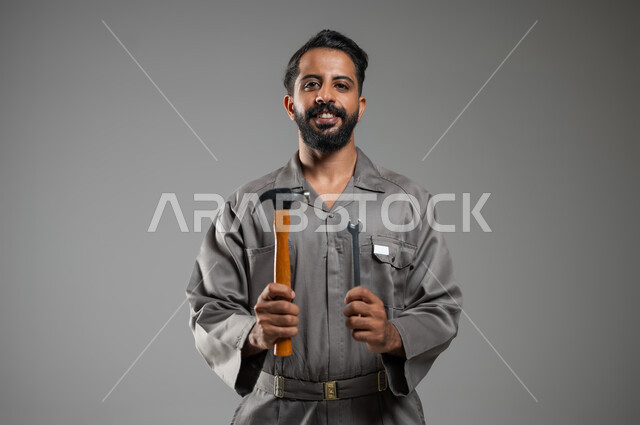 Looking at the camera with pleased gestures, engineering project management, close-up portrait of a Saudi Gulf Arab man wearing a work protective jacket holding a hammer and a wrench in his hands, gray background, sensitive content, sensitive use,