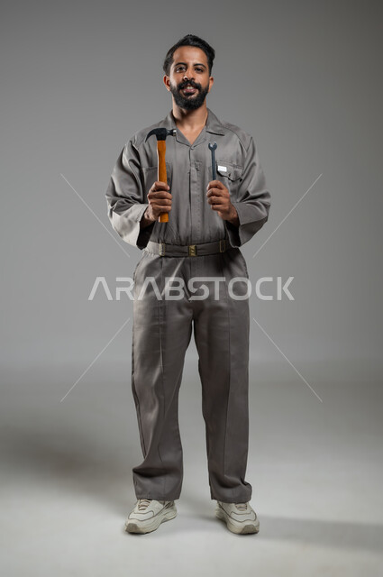 استخدام أدوات ومعدات الصيانة، فني تصليح مبتسم ينظر إلى الكاميرا بإيماءات السرور، المهارة و اتقان العمل، بورتريه لعامل صيانة عربي سعودي خليجي يرتدي يونيفورم العمل يحمل مفتاح الربط والمطرقة، الجسم بالطول الكامل، خلفية رمادية, محتوى حساس, استخدام حساس