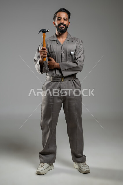 ادارة المشاريع الهندسية، فني تصليح مبتسم ينظر إلى الكاميرا بايماءات السرور، بورتريه لعامل صيانة عربي خليجي سعودي يرتدي سترة حماية خاصة بالعمل يمسك بيده مطرقة,  الجسم بالطول الكامل، خلفية رمادية , محتوى حساس, استخدام حساس