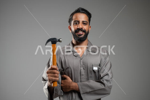 فني تصليح  مبتسم ينظر إلى الكاميرا بايماءات السرور, ادارة المشاريع الهندسية، بورتريه مقرب لعامل صيانة عربي خليجي سعودي يرتدي سترة حماية خاصة بالعمل يمسك بيده مطرقة, خلفية رمادية , محتوى حساس, استخدام حساس