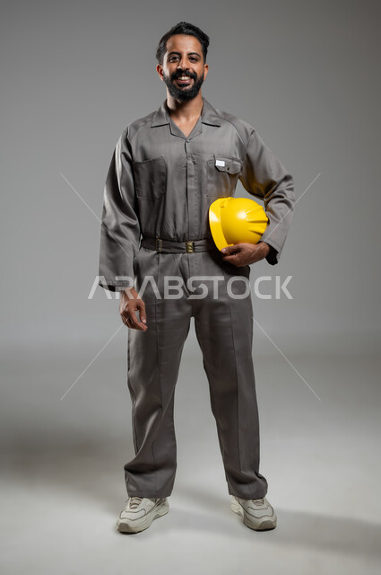 ادارة المشاريع الهندسية، النظر إلى الكاميرا بايماءات السرور، بورتريه بالطول الكامل لمهندس عربي خليجي سعودي يرتدي اليونيفورم الخاص بالعمل يمسك بيده خوذة السلامة, خلفية رمادية,  محتوى حساس, استخدام حساس