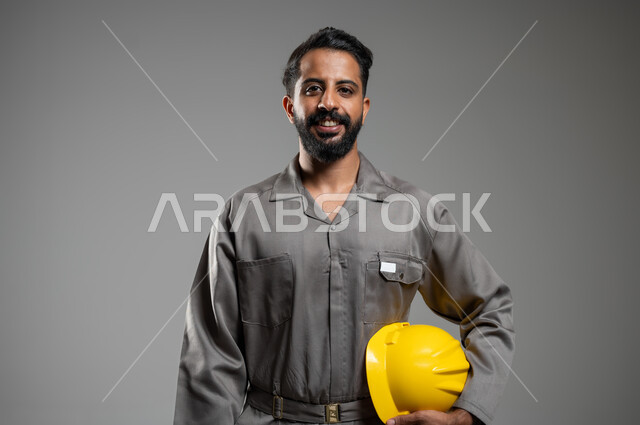 النظر إلى الكاميرا بايماءات السرور ,مهن و وظائف هندسية، بورتريه مقرب لمهندس عربي خليجي سعودي يرتدي سترة حماية خاصة بالعمل يمسك بيده خوذة السلامة, ادارة المشاريع الهندسية، خلفية رمادية, محتوى حساس, استخدام حساس,