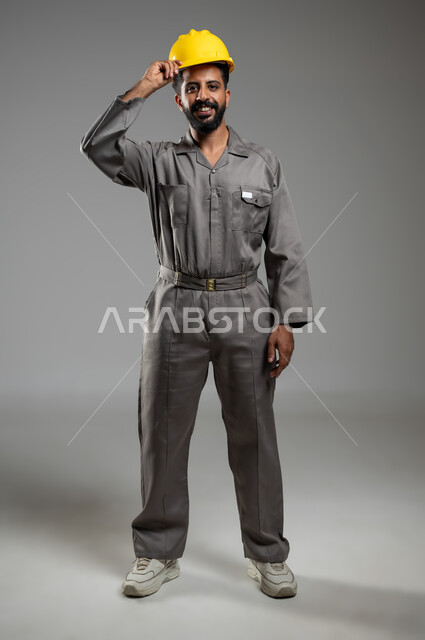 النظر إلى الكاميرا بتعابير السرور، بورتريه لمهندس عربي خليجي سعودي مبتسم يقوم برفع خوذة الحماية احتراما لمهنة الهندسة، مفهوم الانشاءات المعمارية الهندسية، الجسم بالطول الكامل، خلفية رمادية, محتوى حساس, استخدام حساس