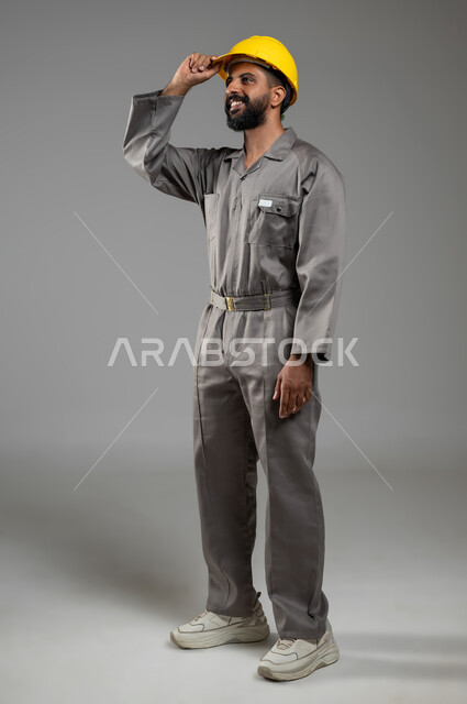 مفهوم الهندسة والانشاءات المعمارية، النظر الى شيء ما بايماءات السرور، مهن ووظائف سعودية، بورتريه لمهندس عربي خليجي سعودي مبتسم يرتدي اللباس الخاص بالعمل يمسك بيده خوذة الحماية بتعابير الفخر، الجسم بالطول الكامل، خلفية رمادية, محتوى حساس, استخدام حساس