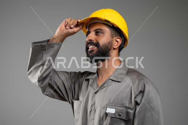 النظر الى شيء ما بايماءات السرور، مهن ووظائف سعودية، بورتريه مقرب لمهندس عربي خليجي سعودي مبتسم يرتدي اللباس الخاص بالعمل يمسك بيده خوذة الحماية بتعابير الفخر، مفهوم الهندسة والانشاءات المعمارية، خلفية رمادية, محتوى حساس, استخدام حساس