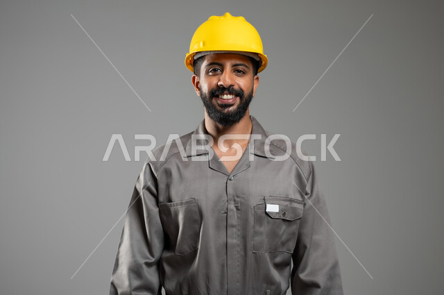 التطور والتقدم في المجال الهندسي، الفخر بالمهن والوظائف السعودية، بورتريه مقرب لمهندس عربي سعودي خليجي مبتسم يرتدي الخوذة وسترة الحماية ينظر الى الكاميرا بإيماءات السرور، خلفية رمادية، استخدام حساس، محتوى حساس