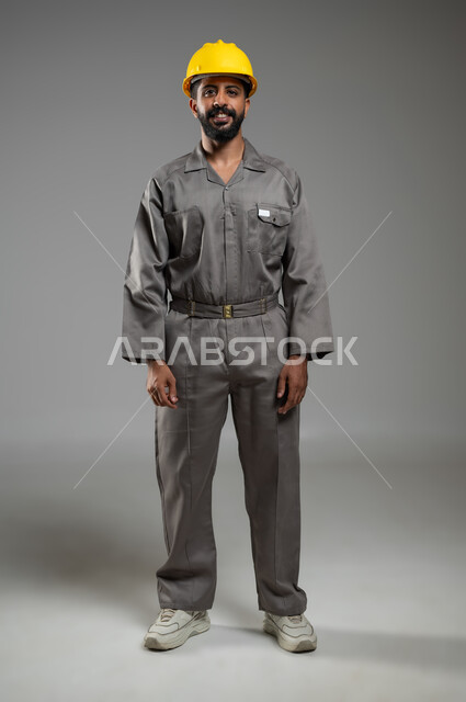 الفخر بالمهن والوظائف السعودية، التطور والتقدم في المجال الهندسي السعودي، بورتريه لمهندس عربي سعودي خليجي مبتسم يرتدي الخوذة وسترة الحماية ينظر الى الكاميرا بإيماءات السرور، الجسم بالطول الكامل، خلفية رمادية، استخدام حساس، محتوى حساس
