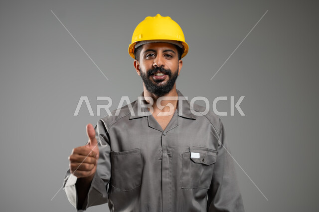Progress in the Saudi engineering field, gestures indicating acceptance and approval, close-up portrait of a Saudi-Gulf Arab engineer wearing a helmet and protective vest looking at the camera with expressions of pleasure, gray background, sensitive use, sensitive content