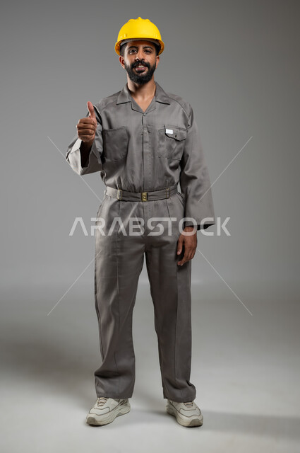 Gestures indicating acceptance and approval, progress in the Saudi engineering field, portrait of a Saudi-Gulf Arab engineer wearing a helmet and protective vest looking at the camera with expressions of pleasure, full-length body, gray background, sensitive use, sensitive content
