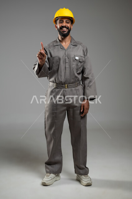 الاشارة بأصبع السبابه الى الأعلى، النظر إلى الكاميرا بايماءات السرور,  بورتريه لمهندس عربي خليجي سعودي يرتدي الخوذة وسترة الحماية يشير باصبعه للاعلى، الجسم بالطول الكامل, خلفية رمادية, محتوى حساس, استخدام حساس