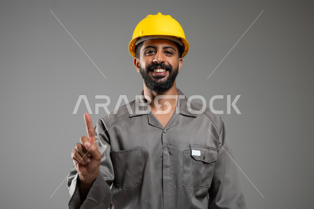 النظر إلى الكاميرا بايماءات السرور, اشارة تمثل الرقم واحد، بورتريه لمهندس عربي خليجي سعودي يرتدي الخوذة وسترة الحماية يرفع اصبع السبابة للأعلى و الاشارة الى شيء ما, الجسم بالطول الكامل، خلفية رمادية, محتوى حساس, استخدام حساس