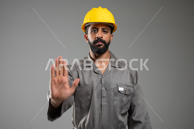 النظر الى الكاميرا ورفع اليد للأعلى للحذر من الاقتراب, بورتريه مقرب لمهندس عربي خليجي سعودي يرتدي الخوذة وسترة الحماية يشير بيده لطلب التوقف والابتعاد,  الامتناع عن فعل أمر معين خلفية رمادية, محتوى حساس, استخدام حساس