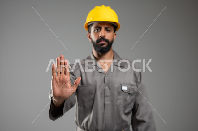 رفع اليد للحذر من الاقتراب, الامتناع عن فعل أمر معين، النظر إلى الكاميرا بايماءات الانزعاج والجدية,  بورتريه مقرب لمهندس عربي خليجي سعودي يرتدي الخوذة وسترة الحماية يشير بيده لطلب التوقف والابتعاد, خلفية رمادية, محتوى حساس, استخدام حساس