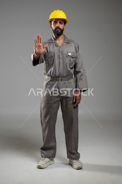 الامتناع عن فعل أمر معين، النظر إلى الكاميرا بايماءات الانزعاج والجدية, رفع اليد للحذر من الاقتراب, بورتريه لمهندس عربي خليجي سعودي يرتدي الخوذة وسترة الحماية يشير بيده لطلب التوقف والابتعاد, الجسم بالطول الكامل, خلفية رمادية, محتوى حساس, استخدام حساس