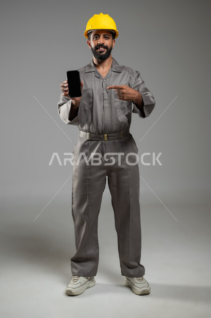 دمج التقنيات الحديثة مع الحياة العملية، النظر إلى الكاميرا بإيماءات السرور، بورتريه لمهندس عربي سعودي خليجي يرتدي الخوذة وسترة الحماية يشير بإصبع السبابة الى شاشة سوداء فارغة عبر الهاتف المحمول،، الجسم بالطول الكامل، خلفية رمادية، استخدام حساس، محتوى حساس