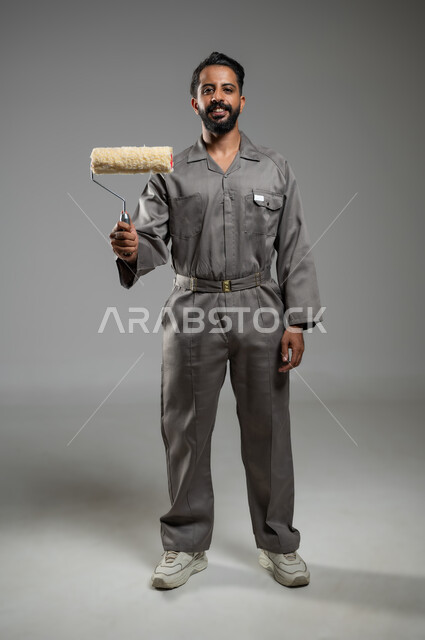 مفهوم المهارة و اتقان العمل، النظر الى الكاميرا بتعابير السرور, مهن ووظائف يدوية سعودية، بورتريه لشاب عربي سعودي خليجي مبتسم يرتدي اليونيفورم الخاص بمهنه البناء والتشييد يحمل بيده بكرة الدهان، الجسم بالطول الكامل, خلفيه رماديه، محتوى حساس، استخدام حساس