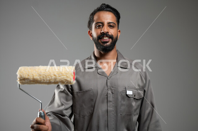 النظر الى الكاميرا بتعابير السرور, مفهوم المهارة و اتقان العمل، مهن ووظائف يدوية سعودية، بورتريه مقرب لشاب عربي سعودي خليجي مبتسم يرتدي اليونيفورم الخاص بمهنه البناء والتشييد يحمل بيده بكرة الدهان، خلفيه رماديه، محتوى حساس، استخدام حساس