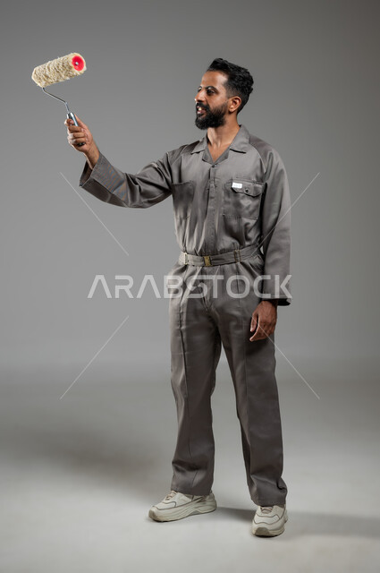 A smiling technical painter doing his work with high efficiency, manual Saudi professions and jobs, a portrait of a Saudi Gulf Arab man wearing the uniform for the building and construction profession, holding a roller in his hand, painting the walls, full-length pictures of the body, gray background, sensitive use, sensitive content