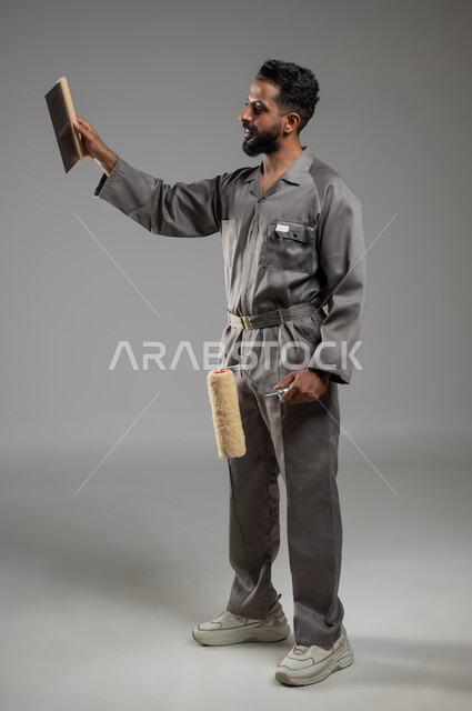 The concept of skill and mastery of work, Saudi manual professions and jobs, a close-up portrait of a smiling Saudi Gulf Arab young man wearing the uniform for the building and construction professions, holding a finishing tool in his hand, practicing his work with high efficiency, full body image, gray background, sensitive content, sensitive use