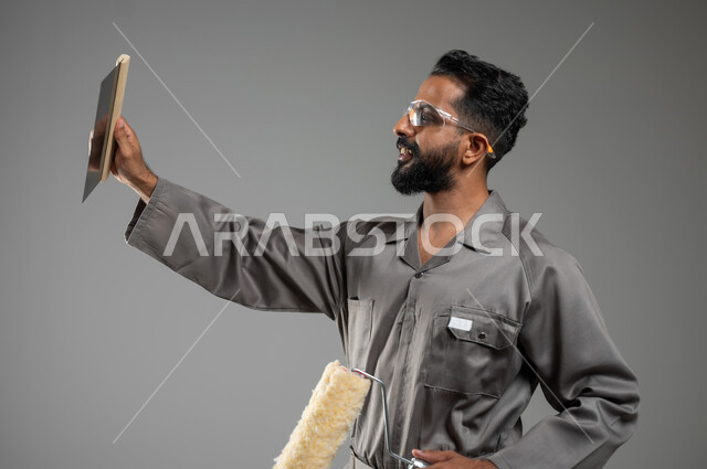 مفهوم المهارة و اتقان العمل، مهن ووظائف يدوية سعودية، بورتريه مقرب لشاب عربي سعودي خليجي مبتسم يرتدي اليونيفورم الخاص بمهنه البناء والتشييد يمسك بيده أدوات الدهان، خلفيه رماديه، محتوى حساس، استخدام حساس