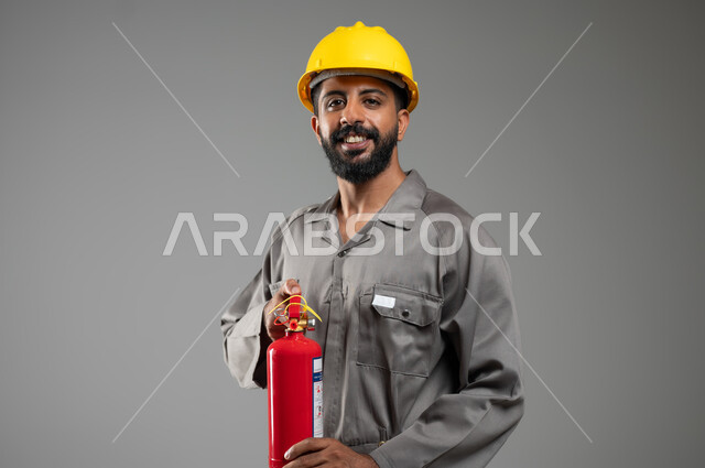 النظر الى الكاميرا بتعابير السرور, مساعدة الناس والتطوع بعمليات الإنقاذ, المعدات اللازمة لإطفاء الحريق ،بورتريه مقرب لرجل عربي خليجي سعودي مبتسم يرتدي سترة الحماية والخوذة يعمل في الدفاع المدني يحمل بيده مطفأة الحريق, خلفية رمادية, محتوى حساس, استخدام حساس