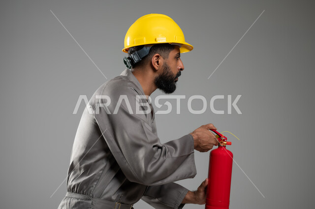 وظائف ومهن سعودية, مساعدة الناس والتطوع بعمليات الإنقاذ, المعدات اللازمة لإطفاء الحريق ،بورتريه مقرب من الجانب لرجل عربي خليجي سعودي يرتدي سترة الحماية والخوذة يعمل في الدفاع المدني يحمل بيده مطفأة الحريق, خلفية رمادية, محتوى حساس, استخدام حساس