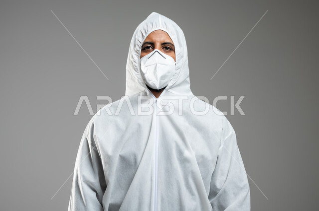 الحفاظ على السلامة و تجنب انتشار العدوى من كوفيد 19 ،الاحترازات الوقائية من فايروس كورونا ، بورتريه مقرب لرجل عربي سعودي خليجي يرتدي الرداء الأبيض البلاستيكي و القناع الطبي ينظر الى الكاميرا , خلفية رمادية, محتوى حساس, استخدام حساس