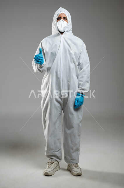 الشعور بالقبول والرضا ,الوقاية من فايروس كورونا، بورتريه لرجل عربي سعودي خليجي يرتدي الرداء البلاستيكي الابيض و القناع الطبي يرفع اصبع الإبهام للأعلى بعلامة الجودة ,تجنب انتشار العدوى من كوفيد 19 ،صورة الجسم بالكامل, خلفية رمادية, استخدام حساس, محتوى حساس