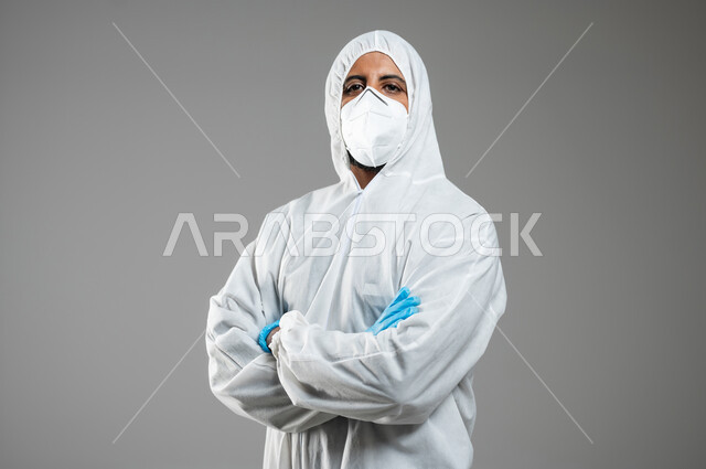 النظر الى الكاميرا بتعابير ايدي متقاطعه, إتباع الاحترازات ضد كوفيد 19، بورتريه مقرب لرجل عربي خليجي سعودي يرتدي الرداء الابيض البلاستيكي و القناع الطبي للوقايه من فايروس كورونا ،خلفيه رمادية, محتوى حساس, استخدام حساس