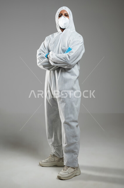 الوقوف بتعابير ايدي متقاطعه, إتباع الاحترازات ضد كوفيد 19، بورتريه لرجل عربي خليجي سعودي يرتدي الرداء الابيض البلاستيكي و القناع الطبي للوقايه من فايروس كورونا ،خلفيه رمادية, صورة الجسم بالكامل, محتوى حساس, استخدام حساس
