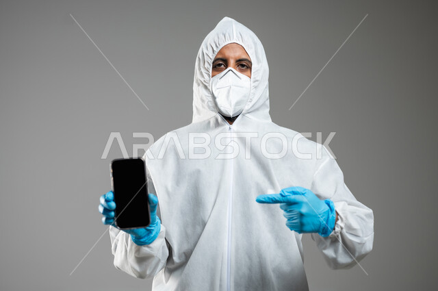 التوعيه من مخاطر كوفيد 19، ارتداء الكمامة لإجراءات الوقاية من فايروس كورونا, بورتريه مقرب لطبيب عربي خليجي سعودي يرتدي رداء أبيض بلاستيكي يشير باصبع السبابة الى الهاتف المحمول بشاشة سودء فارغه، خلفيه رماديه، محتوى حساس، استخدام حساس