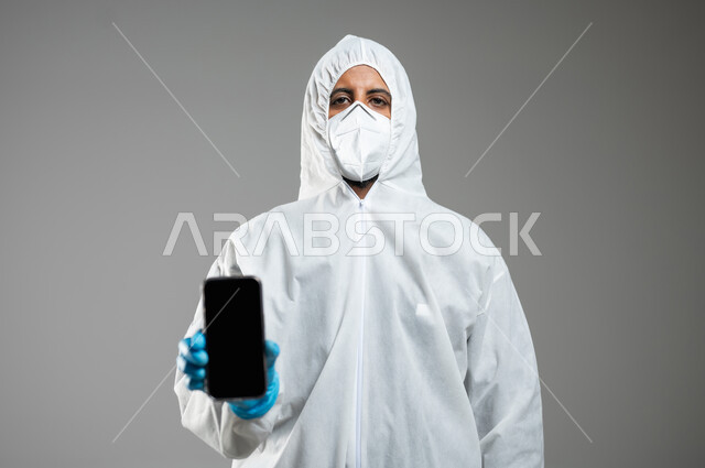 Awareness of the dangers of Covid 19, wearing a mask to prevent the Corona virus, a close-up portrait of a Saudi Gulf Arab doctor wearing a white plastic robe, showing the mobile phone with a blank black screen, gray background, sensitive content, sensitive use