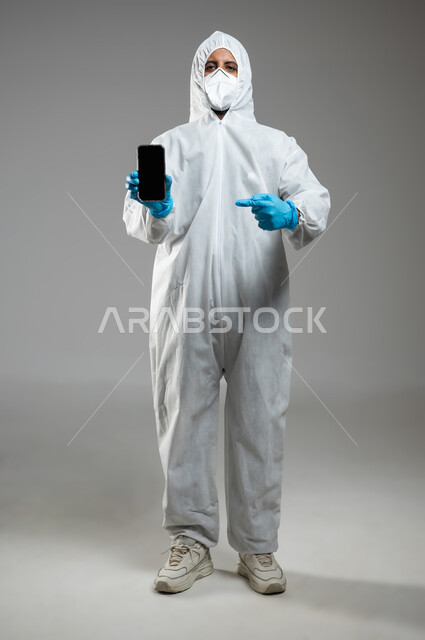 ارتداء الكمامة لإجراءات الوقاية من فايروس كورونا, بورتريه لطبيب عربي خليجي سعودي يرتدي رداء أبيض بلاستيكي  يشير باصبع السبابة الى الهاتف المحمول بشاشة سودء  فارغه، التوعيه من مخاطر كوفيد 19، الجسم بالطول الكامل، خلفيه رماديه، محتوى حساس، استخدام حساس