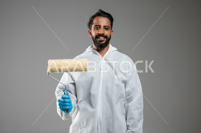 حرف و مهن يدوية، بورتريه مقرب لشاب عربي خليجي سعودي يرتدي اللباس الخاص بالعمل يحمل بيده البكرة يقوم بطلي الجدران، دهان فني مبتسم ينظر الى الكاميرا بتعابير السرور, خلفيه رماديه، استخدام حساس، محتوى حساس