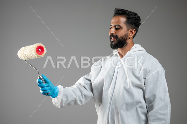 مهن وحرف يدوية، بورتريه مقرب لشاب عربي خليجي سعودي يرتدي اللباس الخاص بالعمل يحمل بيده البكرة يقوم بطلي الجدران، دهان فني مبتسم يمارس مهنته بتعابير السرور, خلفيه رماديه، استخدام حساس، محتوى حساس