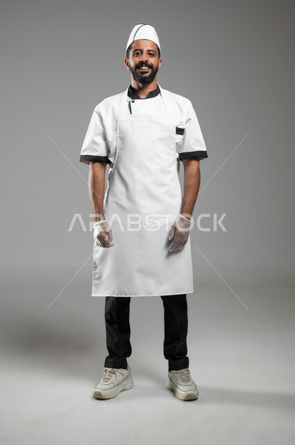 احتراف مهنة الطهي , طهي الطعام السعودي ,بورتريه لشيف عربي خليجي سعودي مبتسم يرتدي يونيفورم الطبخ ينظر إلى الكاميرا بثقة وفخر، صورة الجسم بالطول الكامل, خلفية رماديه، محتوى حساس، استخدام حساس