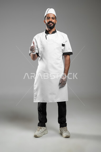 مفهوم اتقان العمل ,طباخ مبتسم ينظر الى الكاميرا بتعابير علامة الجودة ,بورتريه لشيف عربي خليجي سعودي يرتدي يونيفورم الطبخ يرفع اصبع الابهام للاعلى بتعابير الاعجاب، صورة الجسم بالطول الكامل, خلفيه رمادية، محتوى حساس، استخدام حساس