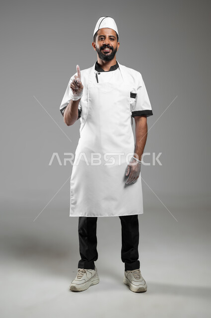 ايماءات الرقم واحد, طباخ عربي خليجي سعودي يرتدي يونيفورم الطبخ ينظر الى الكاميرا بتعابير السرور، بورتريه لشيف يرفع اصبع السبابة للاعلى ويشير الى شيء ما، صورة الجسم بالطول الكامل, خلفيه رماديه، محتوى حساس، استخدام حساس